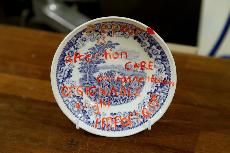 An artwork by Bridget Harvey in the form of a repaired ceramic plate with neon pink writing across it which reads: repair is attention, care, environmentalism, designable, a right, important